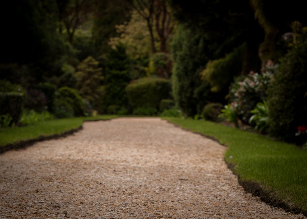 A winding gravel pathway through a lush, green garden with trees and flowering plants on both sides.