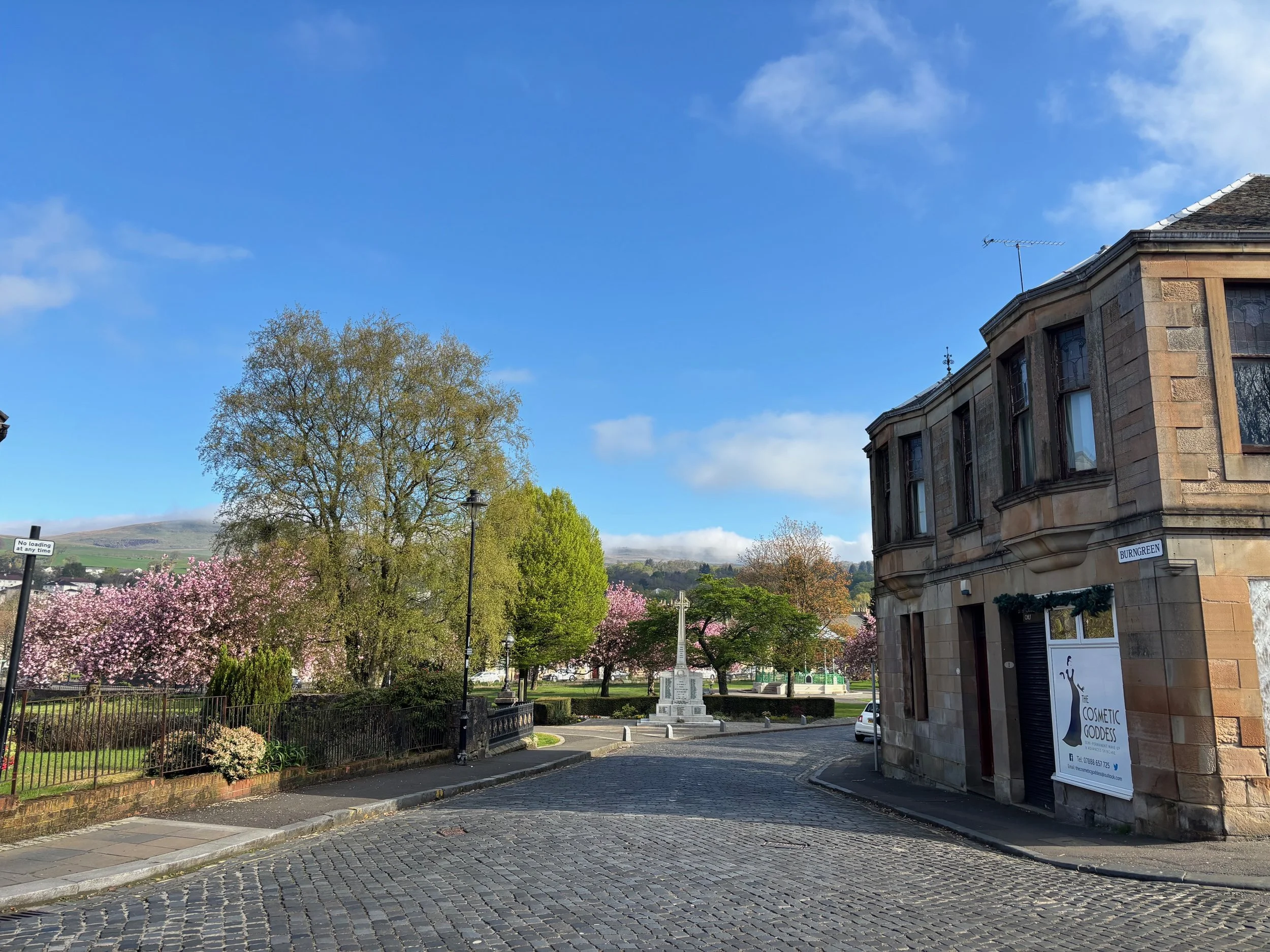 A street scene with cobblestone pavement, a curved building on the right, trees with pink blossoms, a monument or statue in the background, a black lamppost, and a blue sky with some clouds.