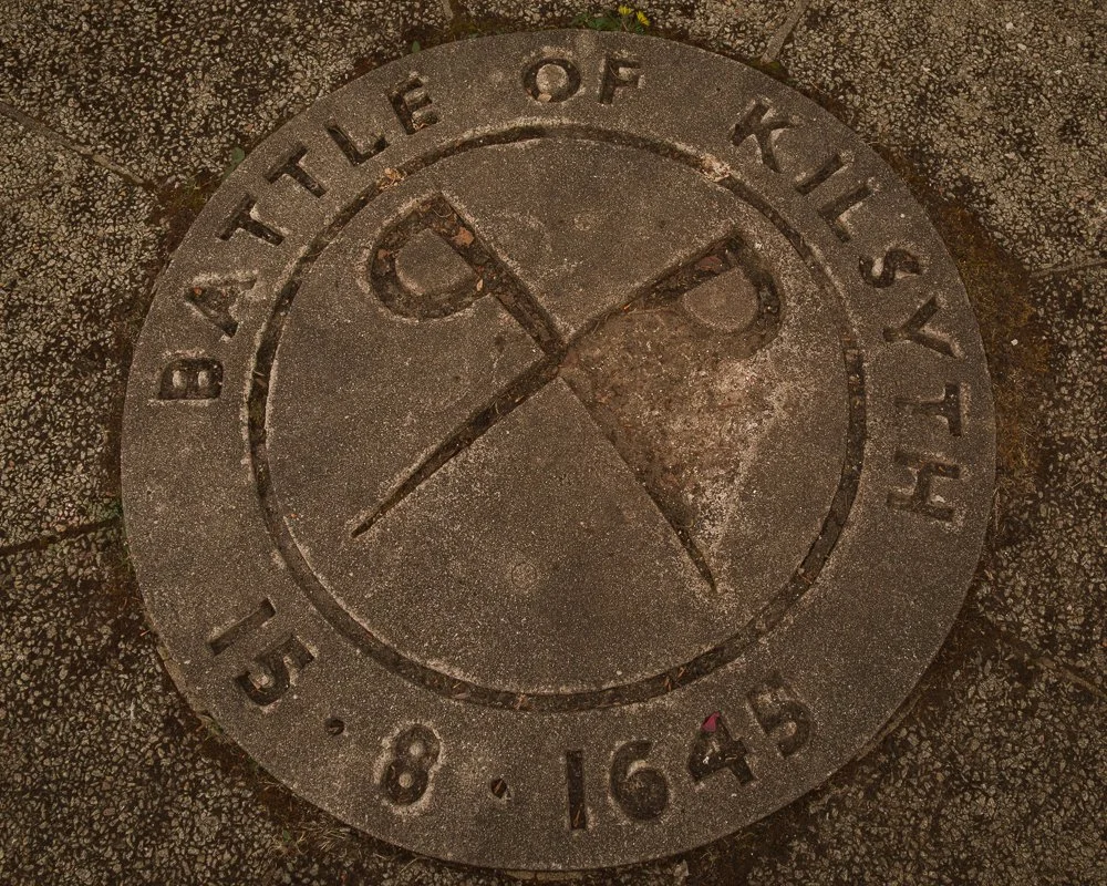 Circular plaque on the ground with the words "The Battle of Kilsyth 15.8.1645" around the edge, featuring a segmented circle with two crossing swords in the center.