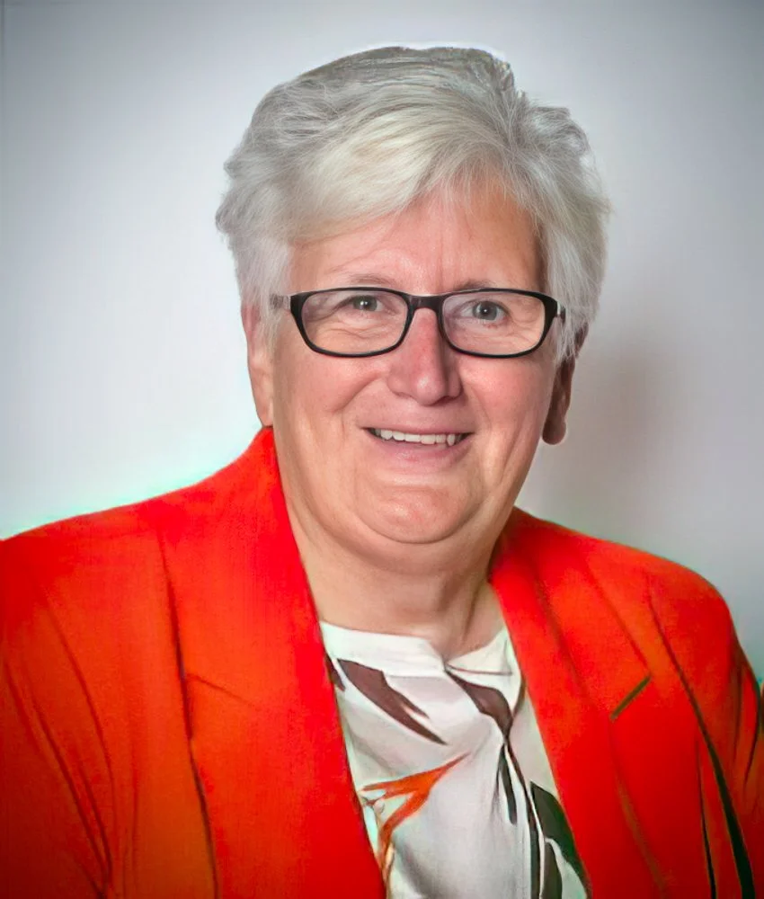 Smiling woman with short gray hair, black glasses, wearing a red blazer and a white blouse with a colorful abstract pattern.