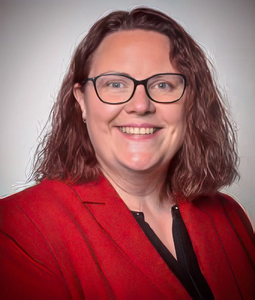 A woman with shoulder-length wavy red hair, wearing glasses, a red blazer, and a black top, smiling at the camera.