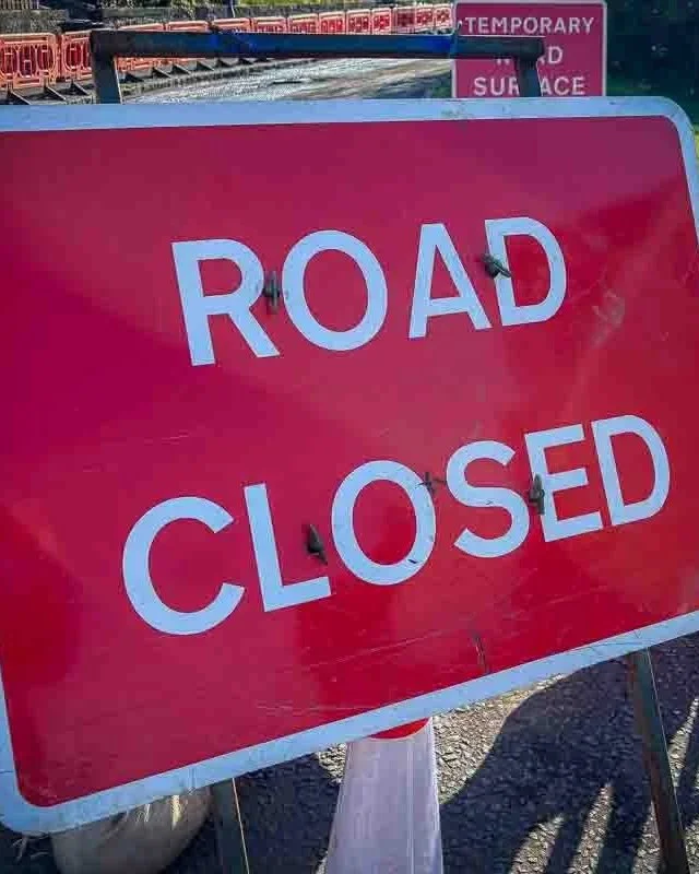 Red road closed sign with white text, placed on a gravel surface with construction barriers visible in the background.