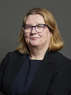 A woman with shoulder-length blonde hair, glasses, wearing a black blazer and a dark top, posing against a plain gray background.