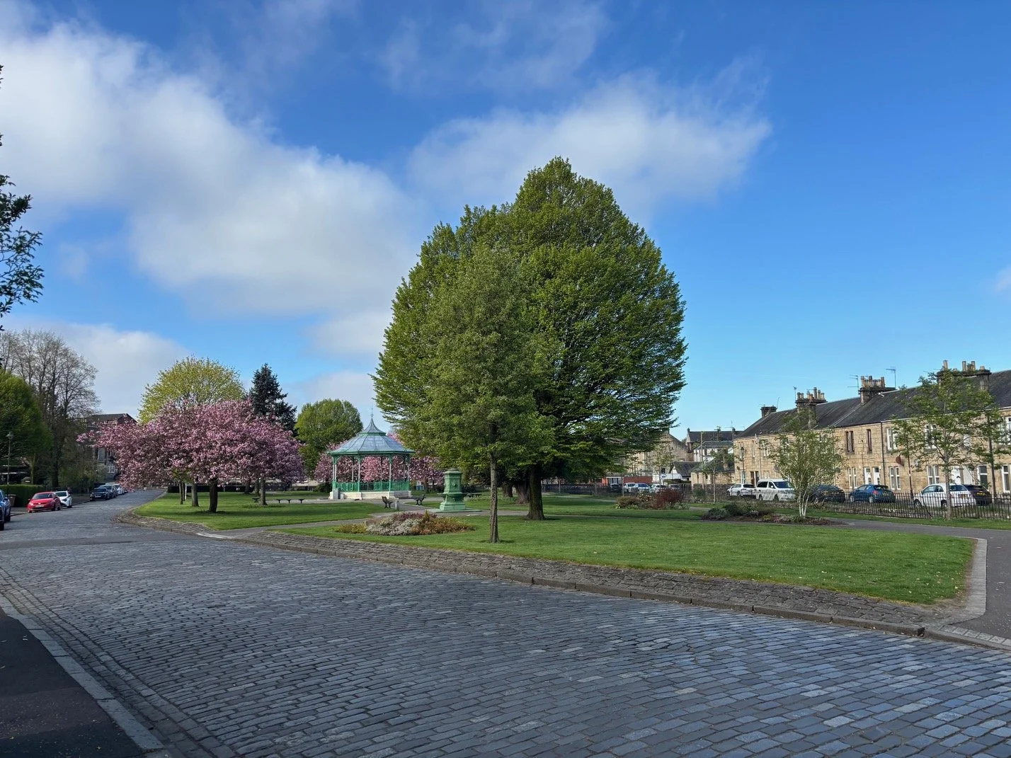 A park with green trees, a pink flowering tree, a gazebo, and a cobblestone walkway under a partly cloudy blue sky, surrounded by residential buildings.