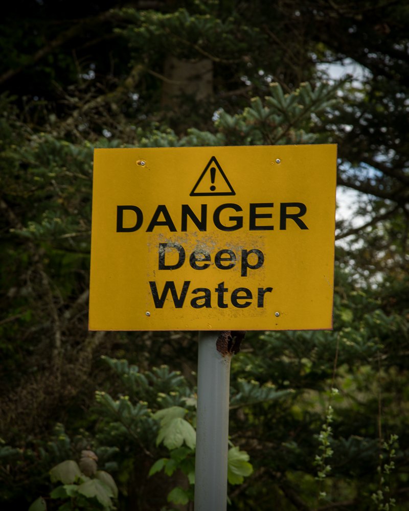 Yellow warning sign with an exclamation mark symbol warning of deep water danger, set against a background of trees.
