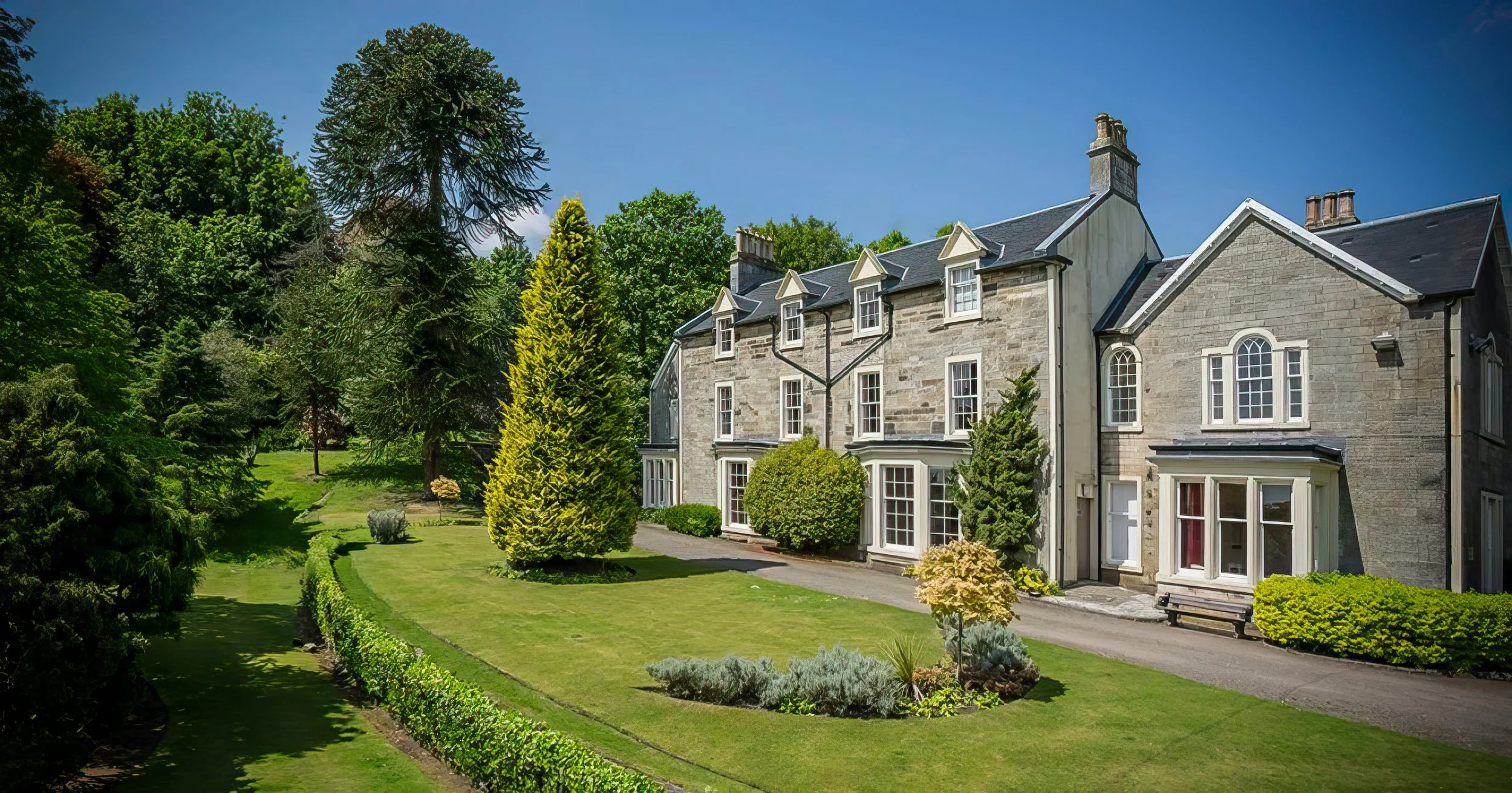 A large stone house with multiple windows and a dark roof, surrounded by landscaped greenery, including trees, bushes, and a lawn under a clear blue sky.