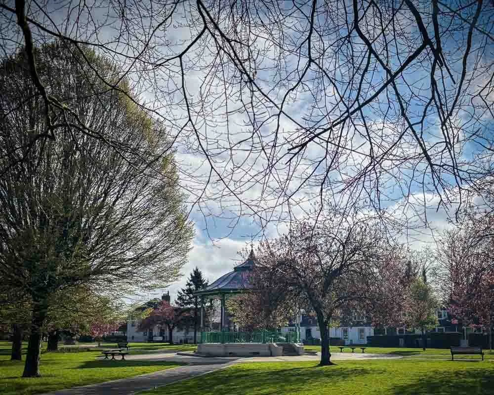 A park with blooming and budding trees, a gazebo, benches, and a walking path beneath a partly cloudy sky.