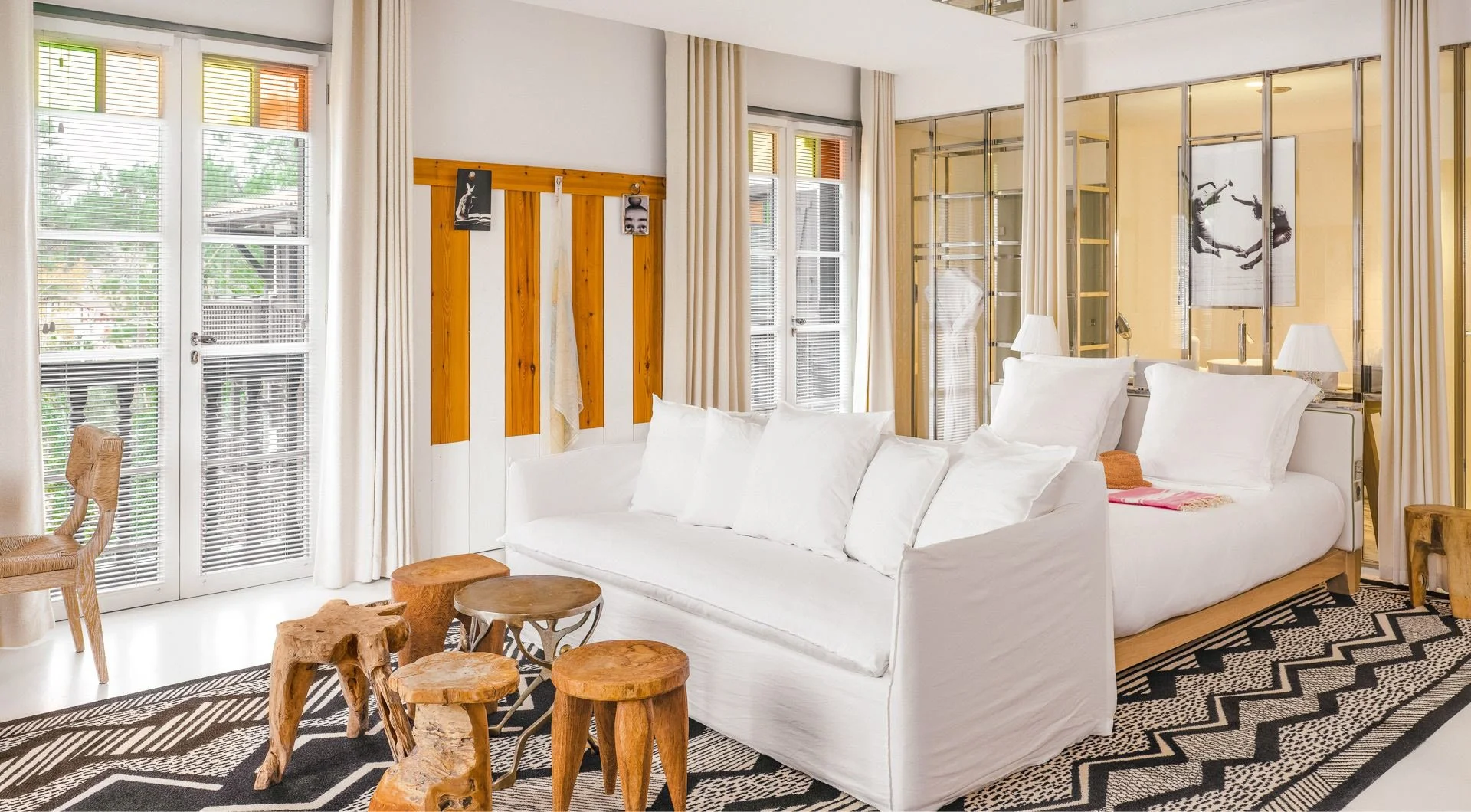 Salon lumineux avec canapé blanc, murs blancs, fenêtres avec volets, décor moderne, tapis noir et blanc, mobilier en bois naturel, divan en rotin, murs séparateurs en verre avec photo d'homme en équilibre. Volets colorés sur les fenêtres.
