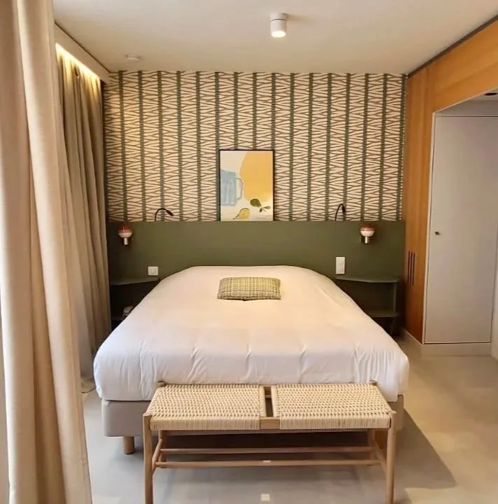 Chambre d'hôtel avec lit double, tête de lit verte, deux lampes de chaque côté, tableau abstrait au mur, et un banc en bois à la base du lit.