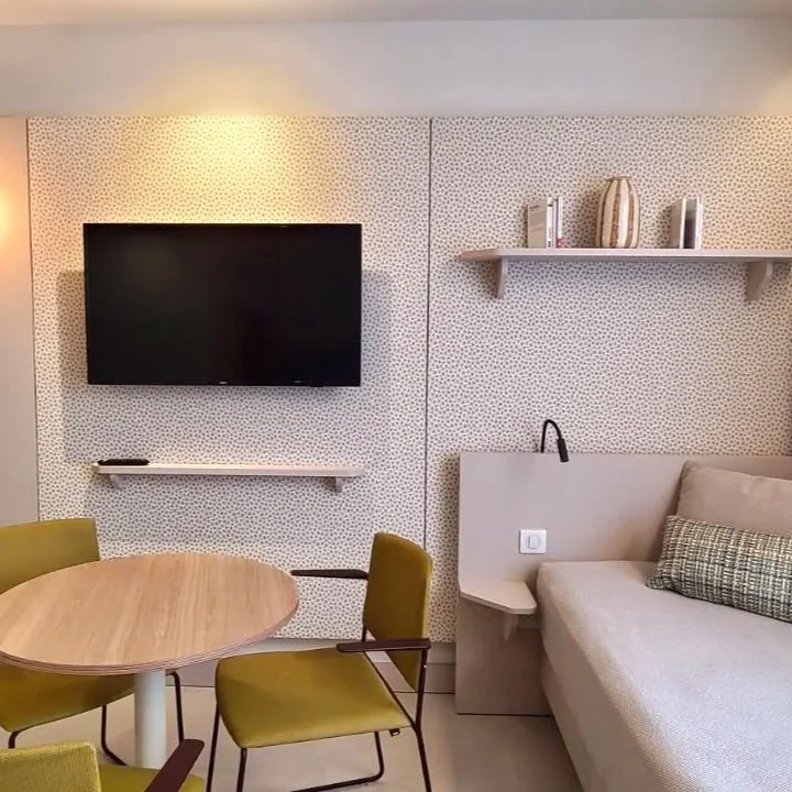 Salon moderne avec télévision à écran plat, canapé beige avec coussin, étagère avec livres et vases, table ronde en bois, quatre chaises vertes