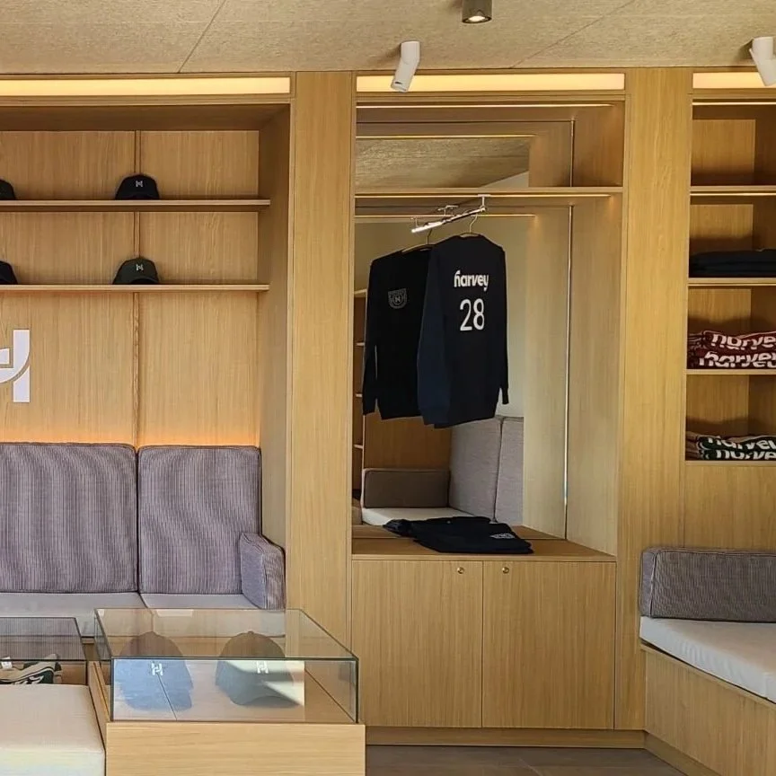 Vitrine en bois contenant des vêtements et des casquettes, avec un banc et un canapé gris en tissu dans une pièce de magasin ou de dressing.