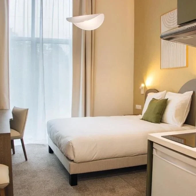 Chambre d'hôtel avec lit double, coussin vert, grande fenêtre avec rideaux, lampe suspendue, petit bureau et chaises.