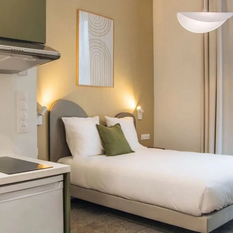 Chambre d'hôtel avec lit double, deux lampes de lecture, tableau moderne au mur, et coin cuisine avec hotte aspirante.
