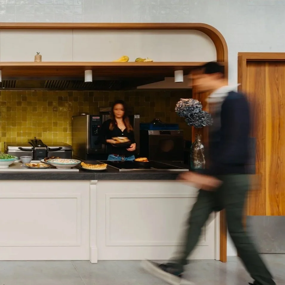 Une cuisine avec deux personnes, une femme derrière le comptoir et un homme en marche devant. Le comptoir est garni de plats de nourriture. La cuisine a un fond de mur avec des carreaux de couleur jaune et des éléments en bois.