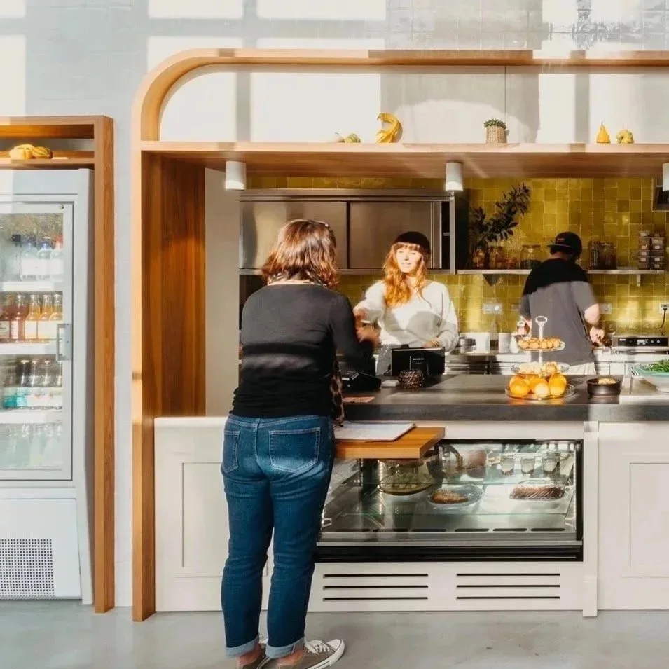 Une femme en train de commander au comptoir d'une petite cuisine ou café, avec deux employés derrière le bar et un réfrigérateur à gauche.