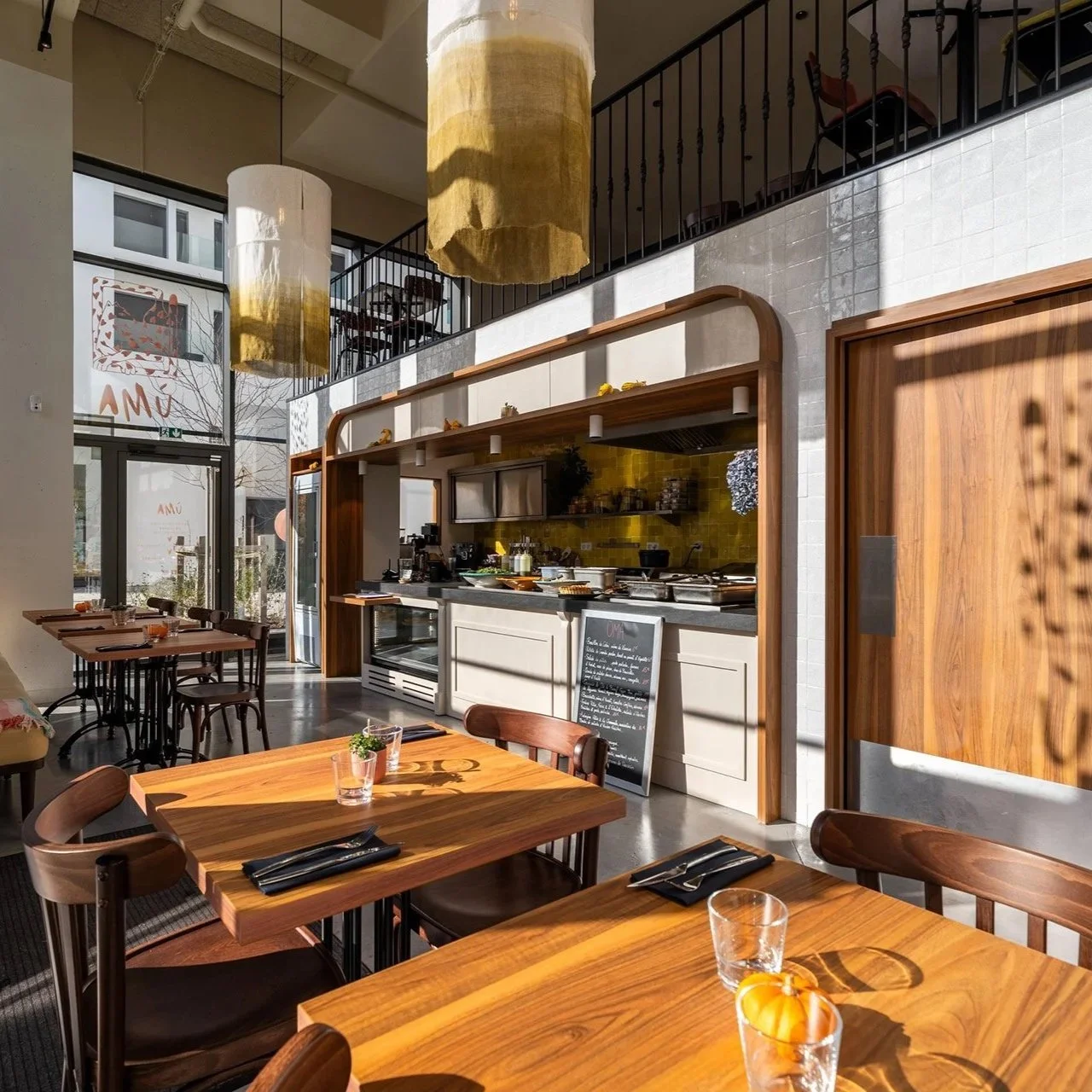 Intérieur d'un café ou restaurant avec tables en bois, vases avec citrouilles, cuisine en arrière-plan avec meubles en bois et murs blanc. Grandes fenêtres laissant entrer la lumière naturelle, éclairage suspendu en haut, mezanine avec chaises.