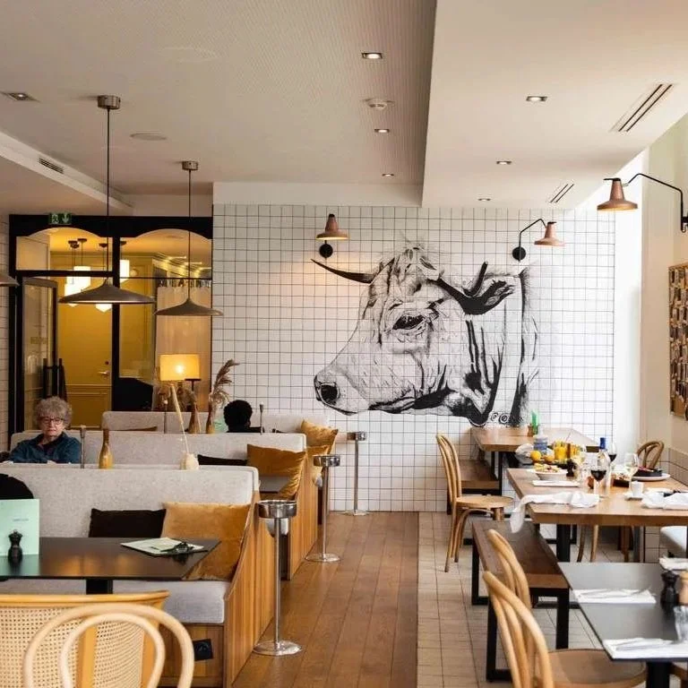 Intérieur d'un restaurant avec un mur décoratif représentant un taureau en style dessin, tables en bois, chaises, lumières suspendues, et quelques clients.