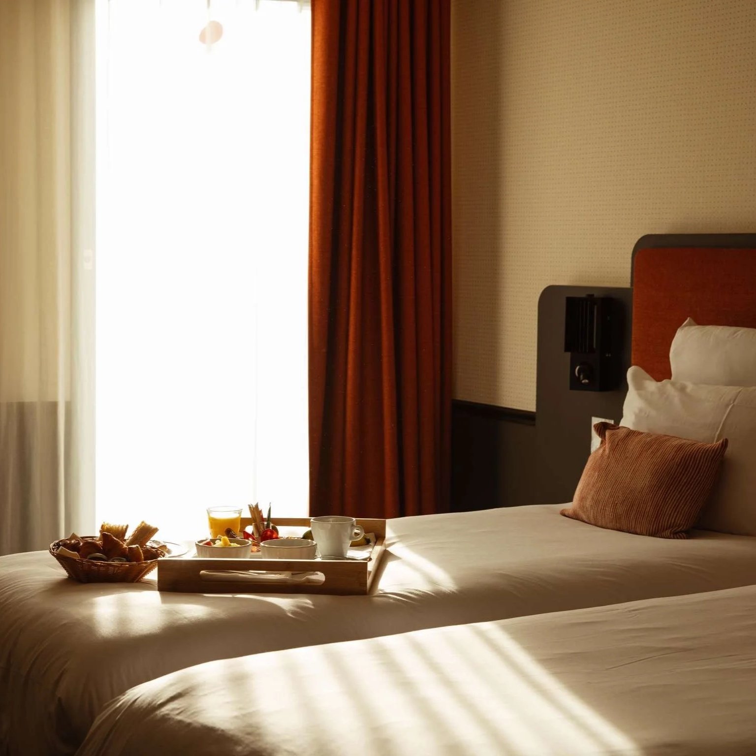 Chambre d'hôtel avec lit fait, petit déjeuner sur un plateau, rideaux marrons, lumière naturelle