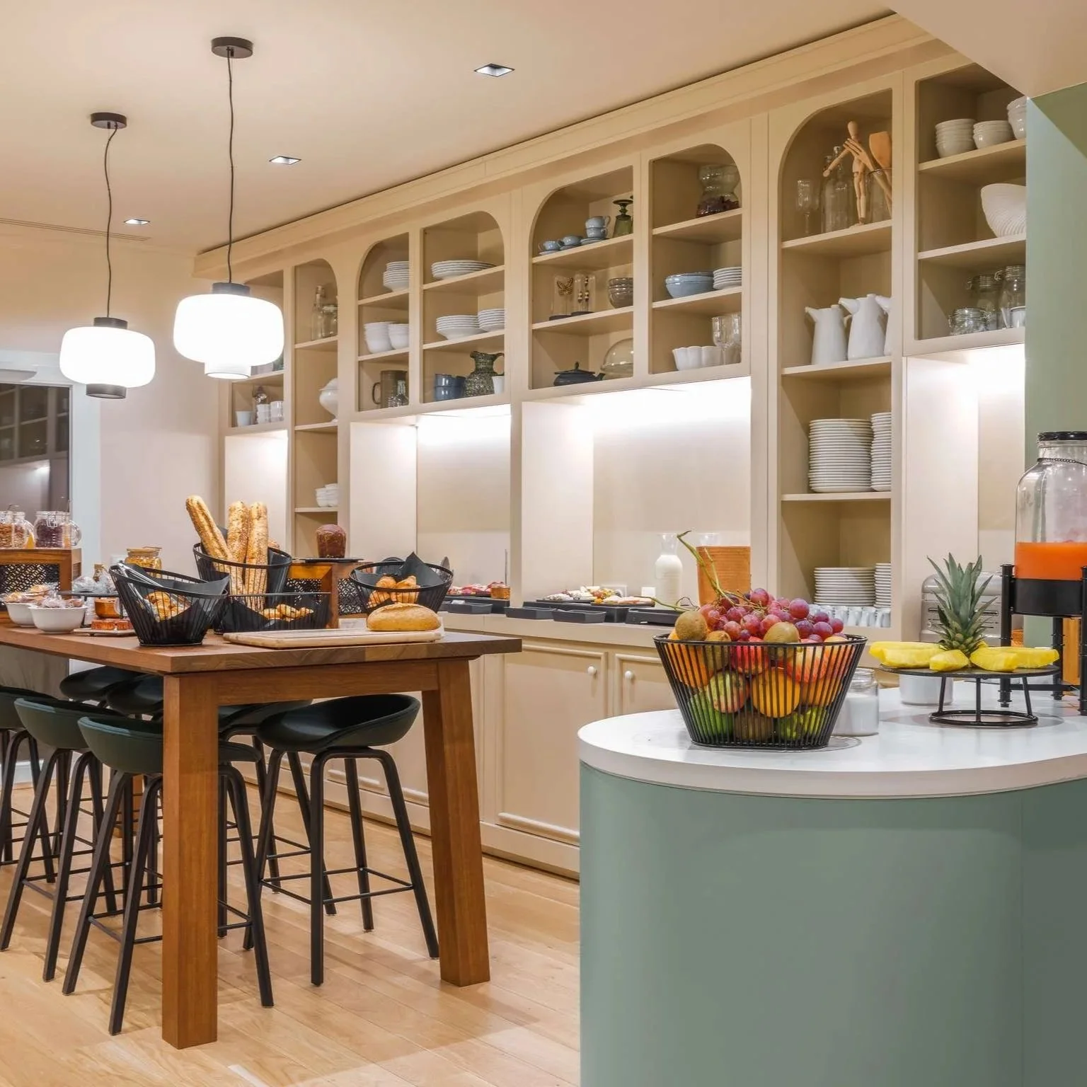 Une cuisine moderne avec un îlot central en bois, des étagères remplies de vaisselle blanche, un buffet avec divers aliments et fruits sur la table, y compris des raisins, une ananas et des bananes, et une lumière chaleureuse.