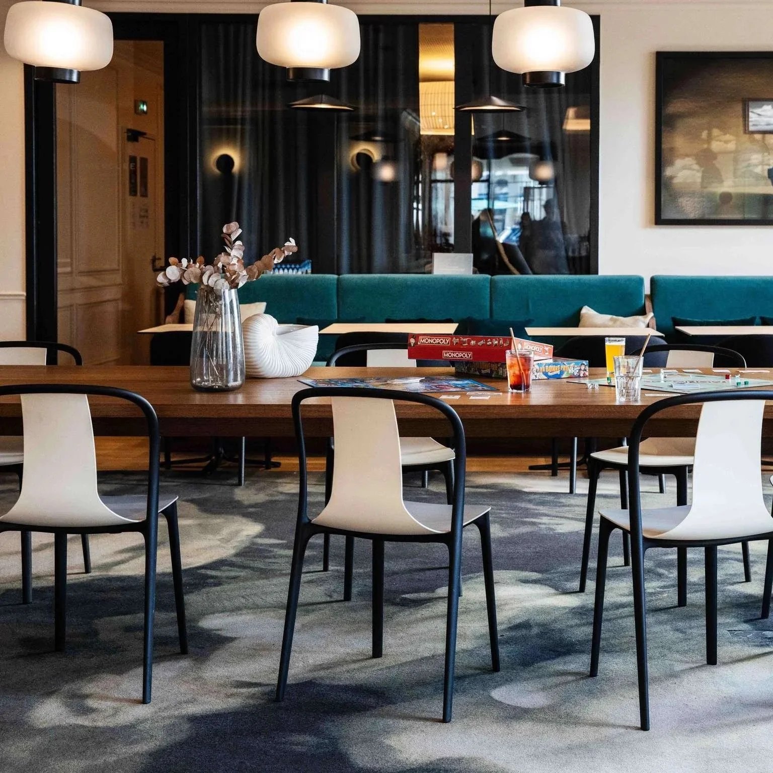Intérieur d'un restaurant moderne avec une grande table en bois, chaises noires et blanches, et un fond avec un canapé vert turquoise, des lampes suspendues au plafond, et une décoration minimaliste avec une vase contenant des branches séchées, des j