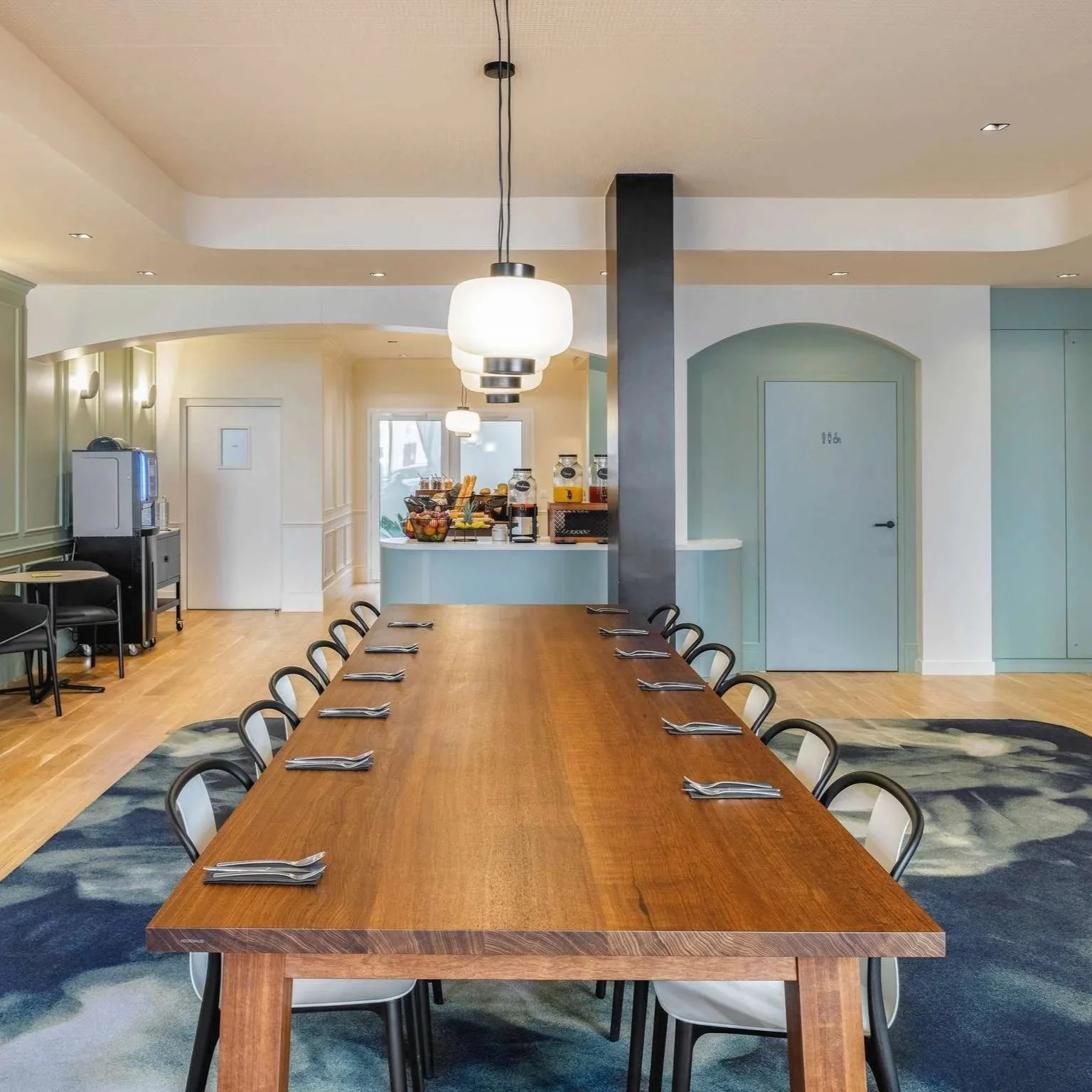 Salle de réunion avec grande table en bois, chaises noires, vaisselle pour chaque place, éclairage suspendu moderne, buffet avec fruits et boissons en arrière-plan, décoration sobre et moderne.