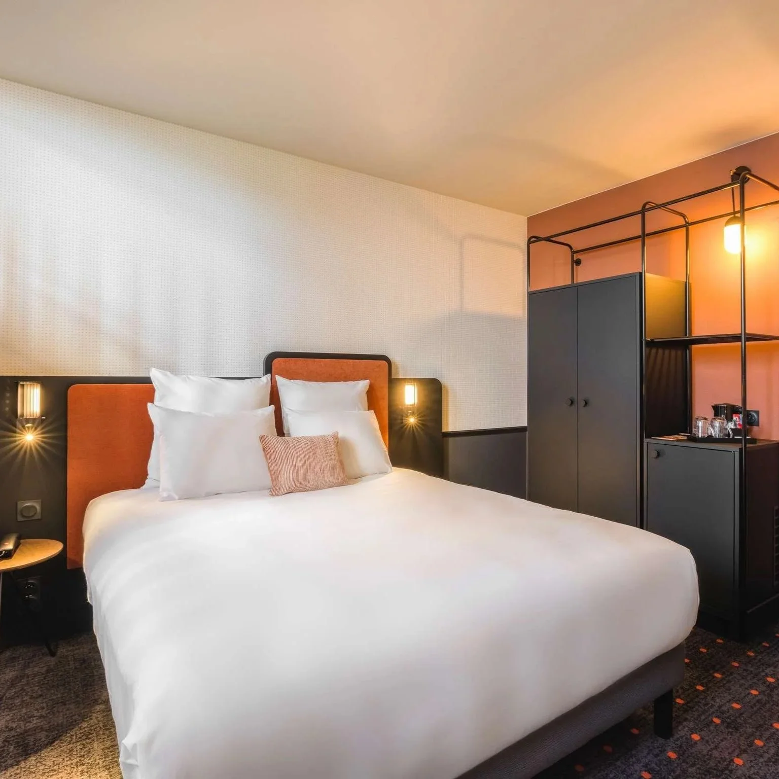 Chambre d'hôtel moderne avec lit blanc, tête de lit orange et meubles noirs, éclairage chaud.