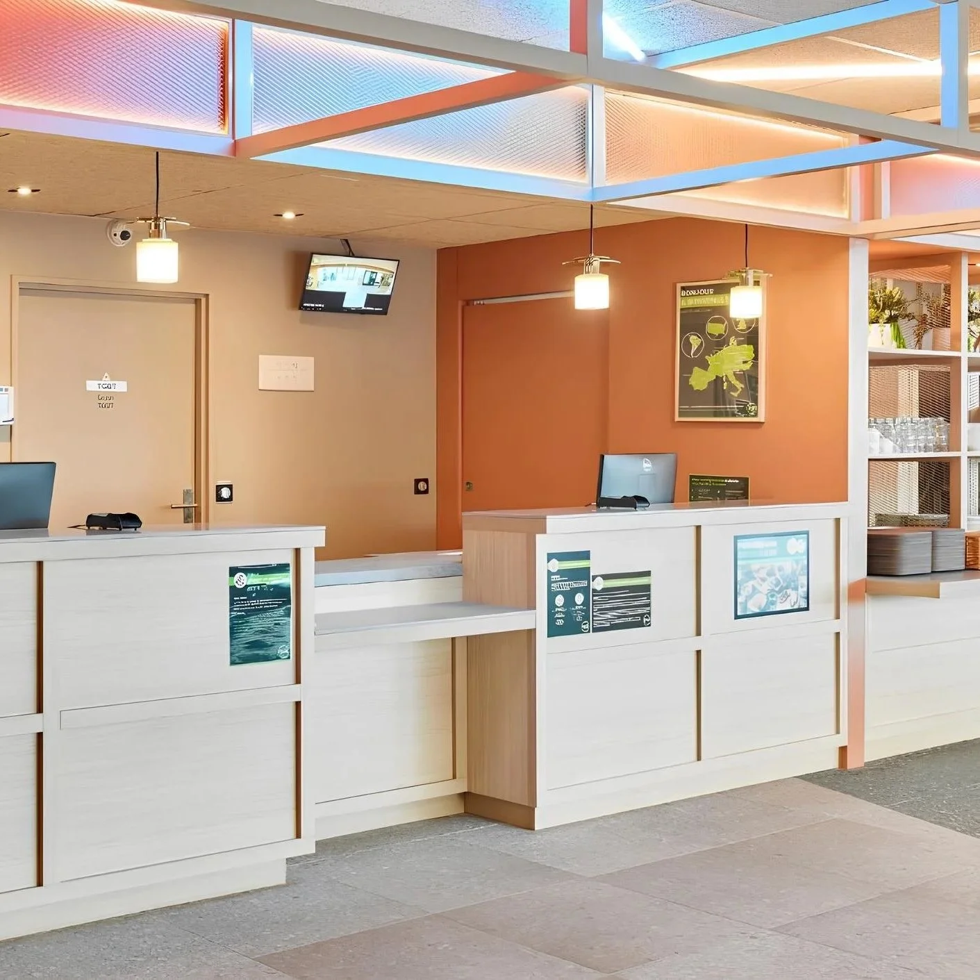 Comptoir d'accueil dans un espace moderne avec une frontière blanche, des écrans, des affiches, des lampes suspendues, et un mur orange avec une affiche.