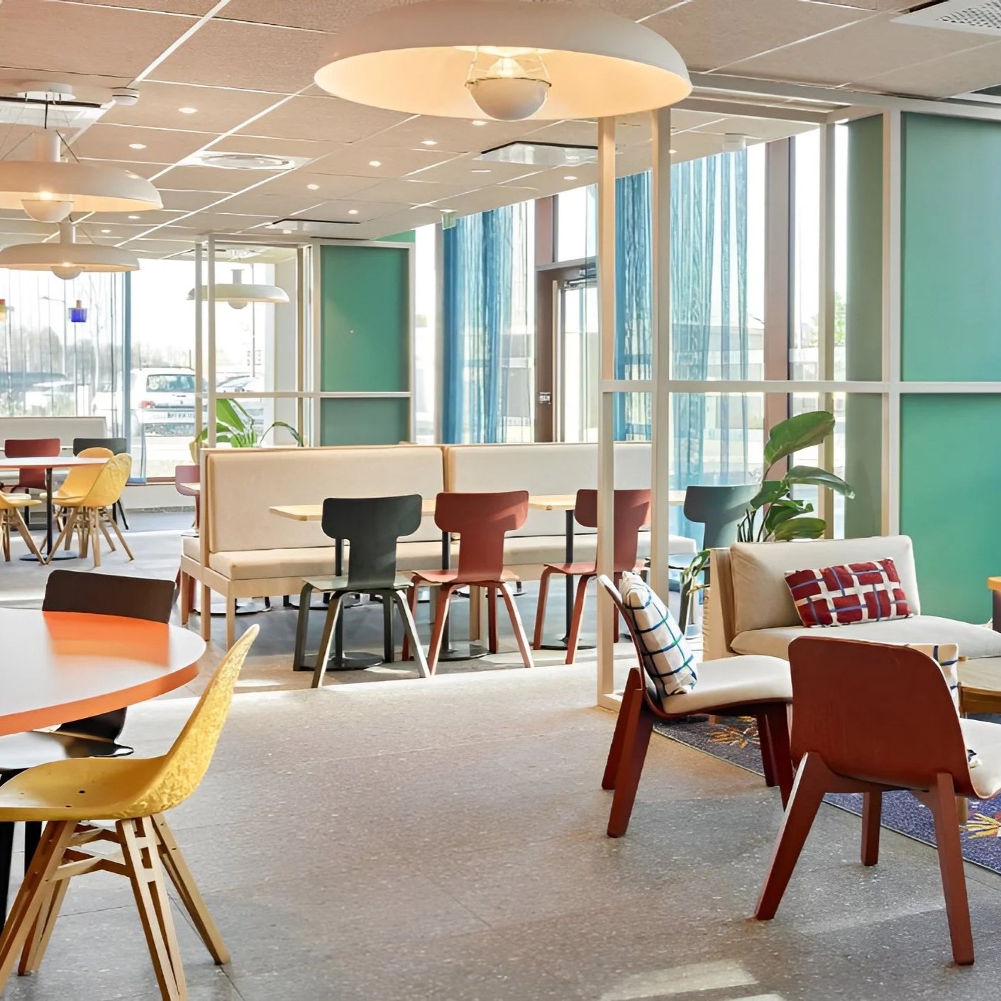 Intérieur d'un café ou restaurant lumineux avec diverses tables et chaises colorées, grands fenêtres avec rideaux bleus et plantes vertes.