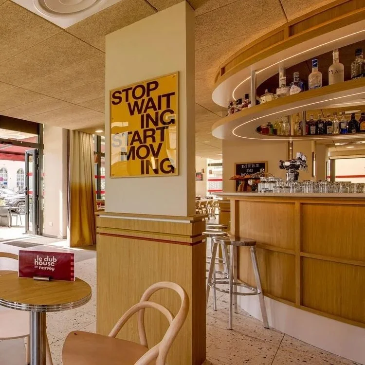 Intérieur d'un bar ou restaurant lumineux avec un comptoir en bois, étagères avec bouteilles d'alcool, et une affiche jaune avec des mots en anglais. La salle est bien éclairée, avec des fenêtres laissant entrer la lumière naturelle.