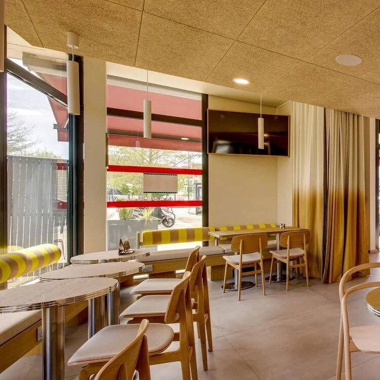 Intérieur d'un café avec de grandes fenêtres, chaises en bois et tables en marbre, un écran plat fixé au mur, et des rideaux beige.