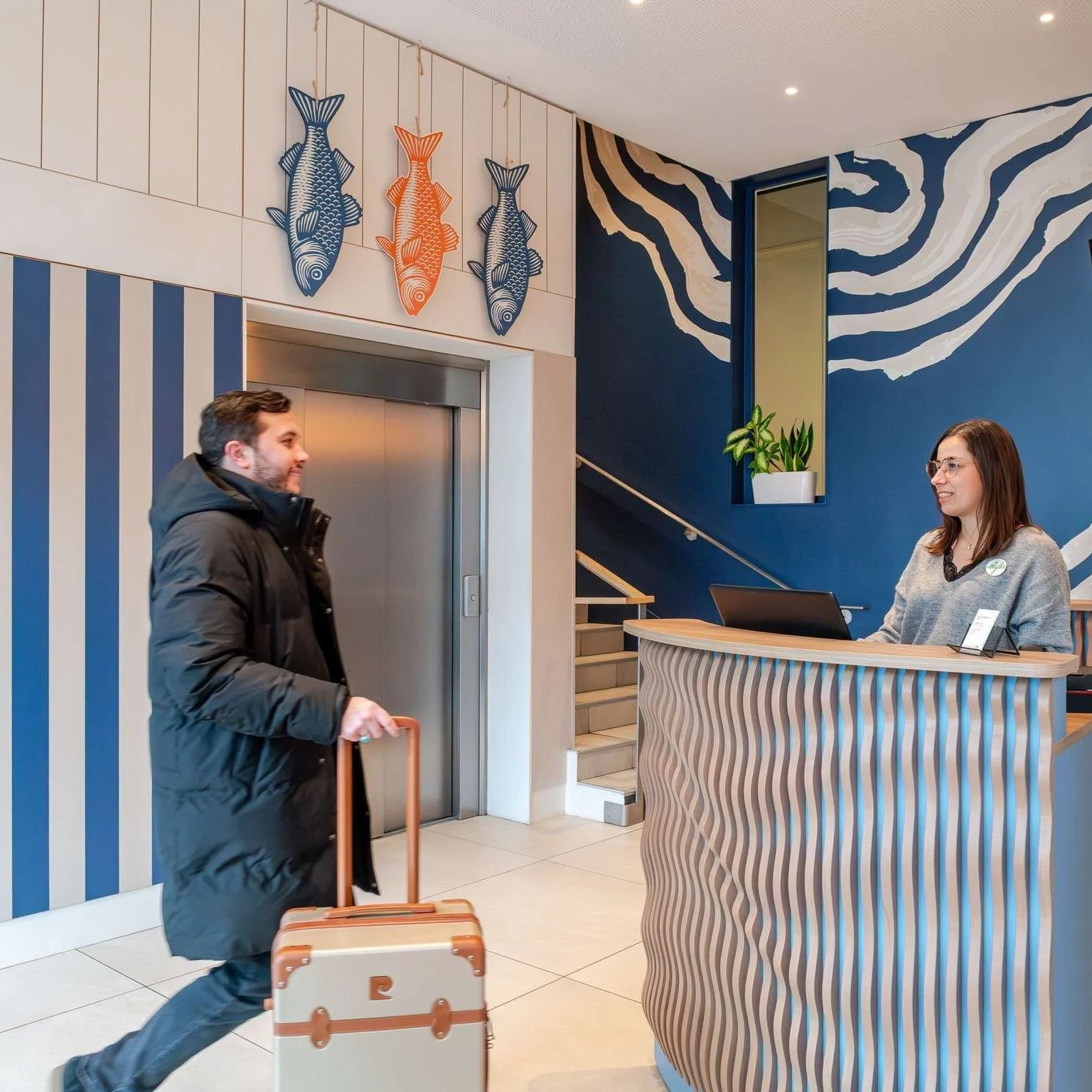 Homme avec valise à roulettes arrivant à la réception d'un hôtel, conversant avec une réceptionniste derrière un comptoir en bois incurvée. Décor intérieur moderne avec mur bleu, sculpture de poissons en haut, et escalier en arrière-plan.