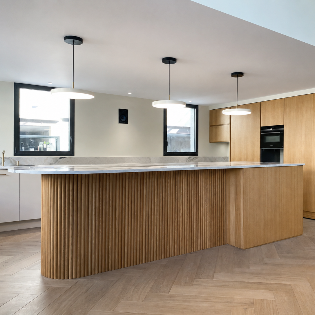 Cuisine moderne avec îlot en bois avec moulures verticales, fenêtres avec vue sur l'extérieur, luminaires suspendus blancs, planche de bois au sol, objets sur le plan de travail comme tasses, sac, et un robot de cuisine.