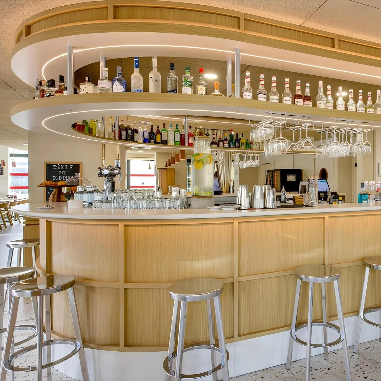 Un bar moderne avec comptoir en bois clair, étagères en haut avec bouteilles d'alcool, verres suspendus, et un distributeur de citronnade glacée.