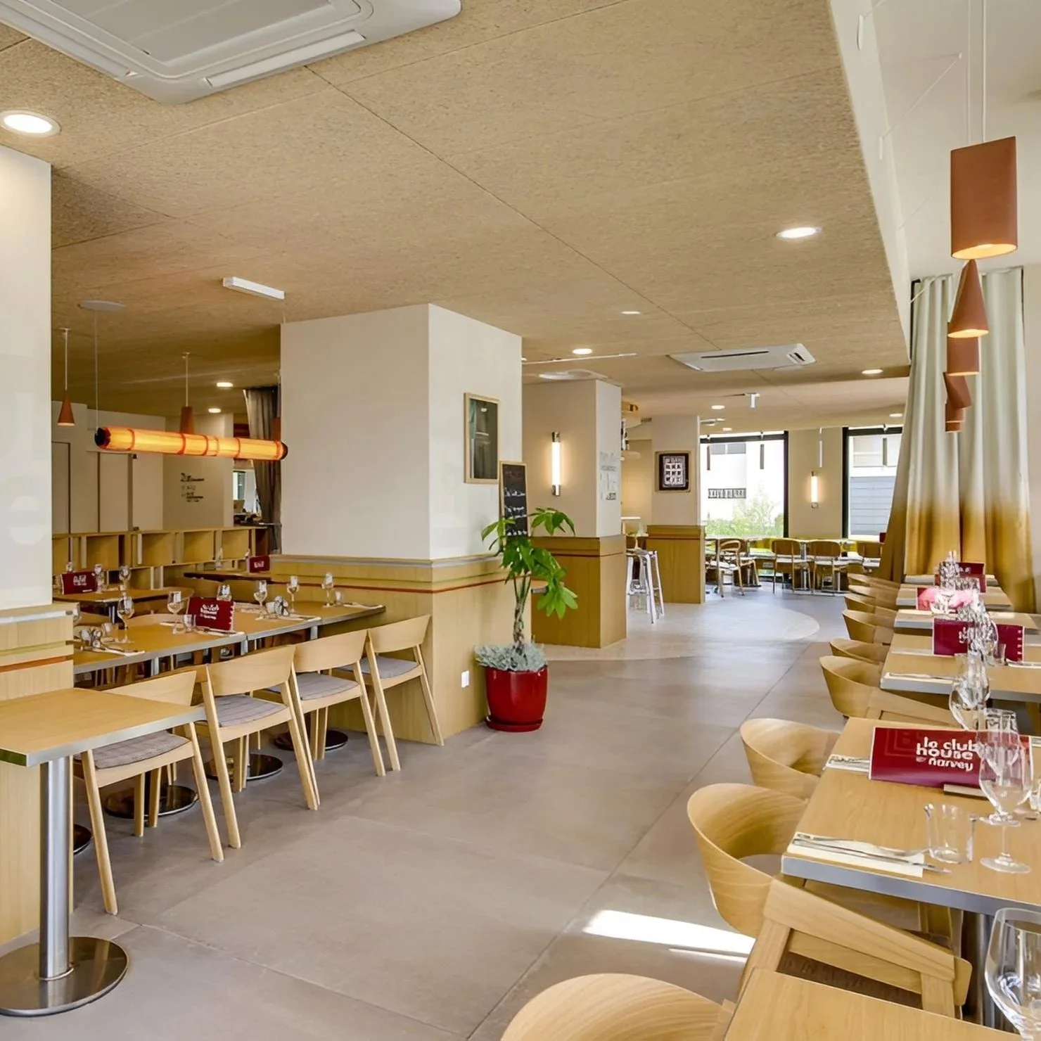 Salle à manger d'un restaurant lumineux avec tables en bois, chaises en bois beige, couverts, verres, nappes rouges, et décor simple.