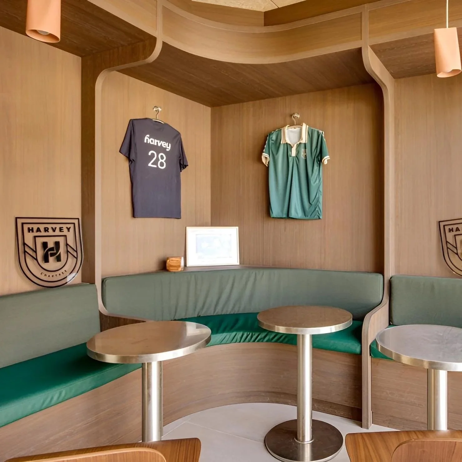 Espace de salon avec mur en bois, deux maillots de sport suspendus, une bannière de football Harvey, un canapé en U avec coussins verts, et deux petites tables rondes en métal.