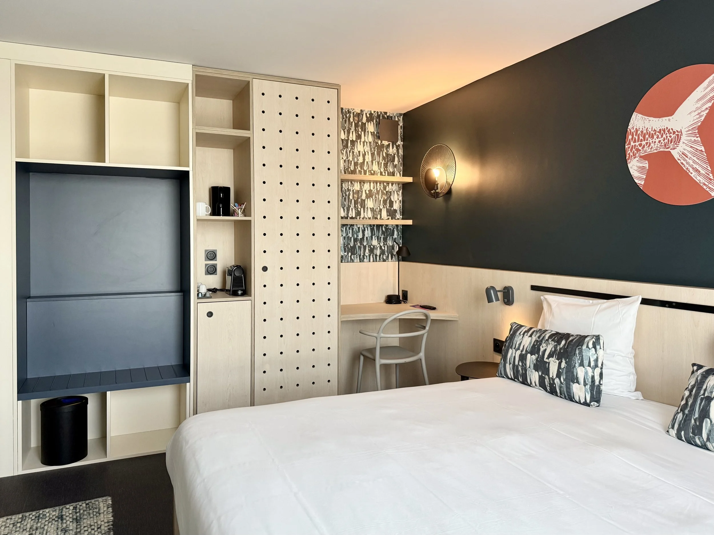 Chambre d'hôtel avec un lit, des oreillers, un meuble avec étagères et une lampe murale. Mur blanc et mur noir avec une décoration en forme de étoile de mer.