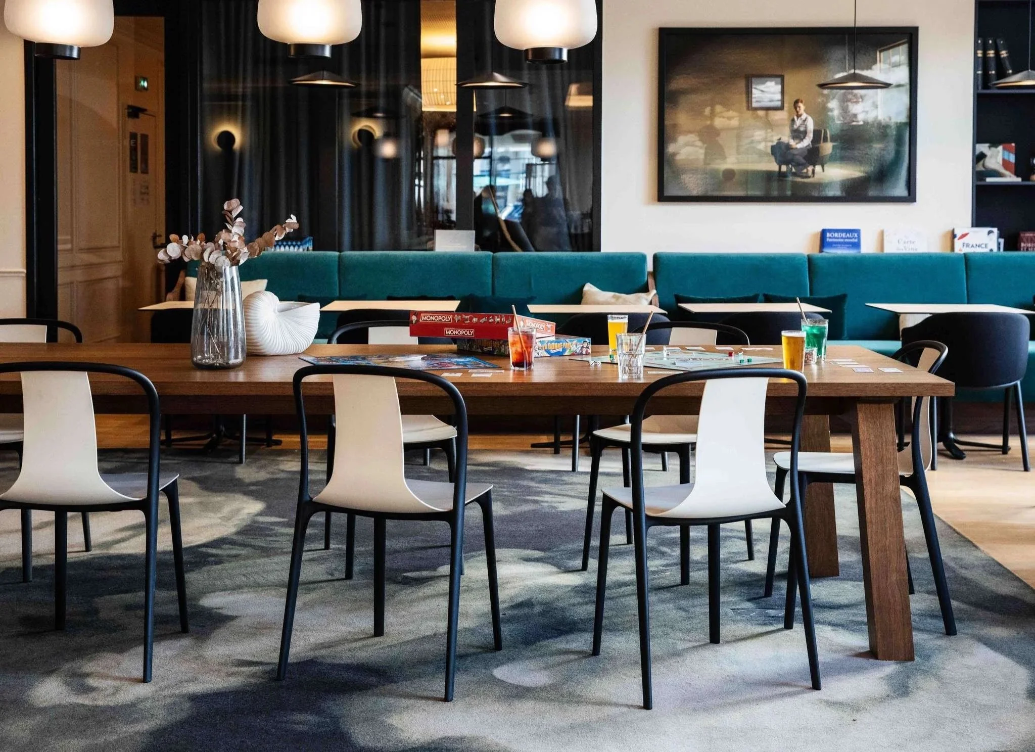 Intérieur d'un café ou restaurant moderne avec des tables, des chaises blanc et noir, une bannette bleue avec des livres et magazines, un vase avec des fleurs en devanture, une grande affiche ou peinture avec une femme assise dans un paysage naturel, des lampes suspendues et un décor épuré.