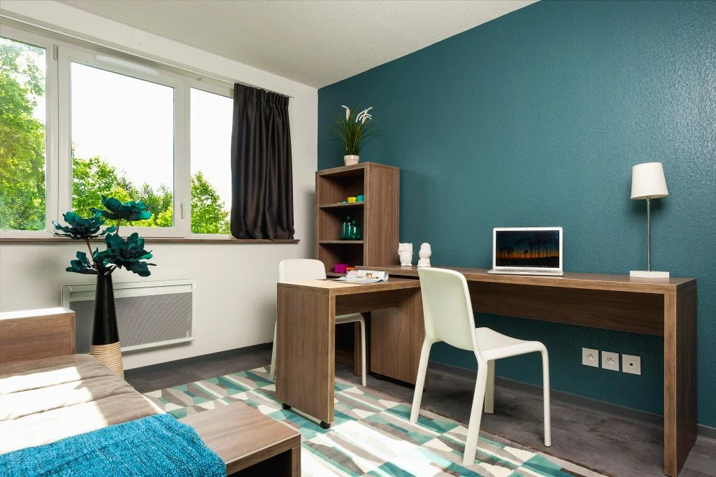 Une chambre avec un bureau en bois, une chaise blanche, une lampe de bureau, une étagère en bois avec des plantes et des décorations, un lit partiellement visible, un tapis à rayures, et une fenêtre donnant sur des arbres verts.