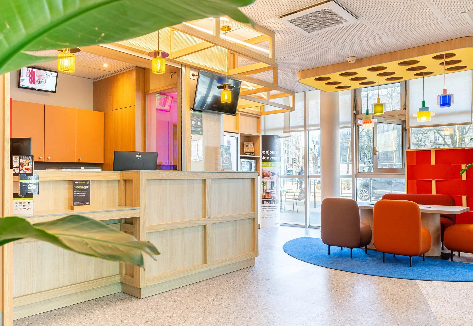 Intérieur d'une salle d'accueil ou de réception avec un comptoir en bois clair, des lampes suspendues jaunes, un mur avec un écran TV, des fenêtres laissant entrer la lumière naturelle, et des fauteuils orange et beige dans un coin salon.