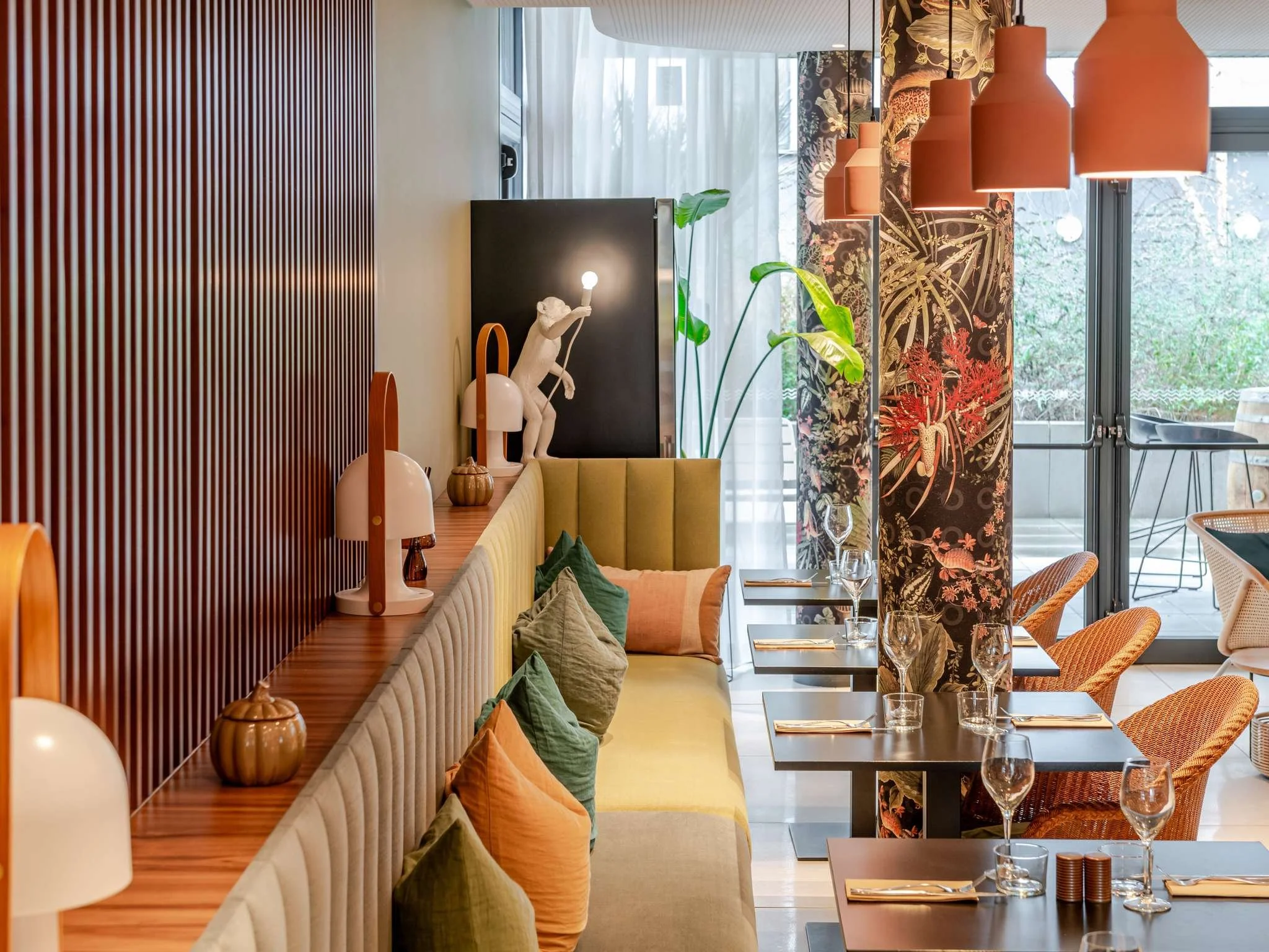 Intérieur d'un restaurant moderne avec une décoration colorée, comprenant des lampes orange, des fauteuils jaune et orange, des plantes vertes, et une salle de restaurant avec des tables dressées pour un repas.
