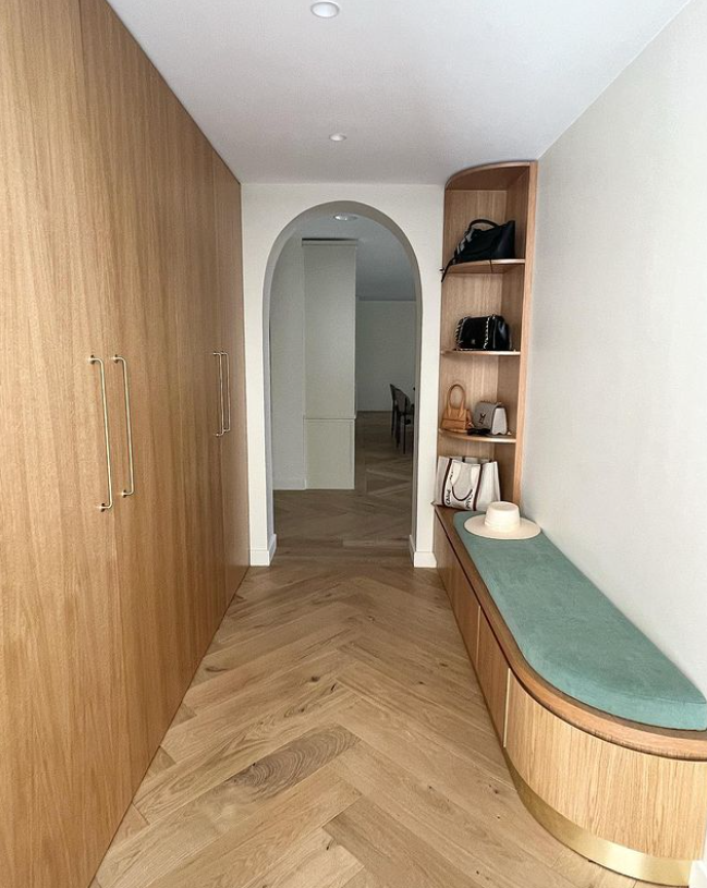 Un couloir avec un placard en bois sur le côté gauche et un banc en bois avec un coussin vert sur la droite, avec des sacs à main et un chapeau posés dessus, et un mur blanc.