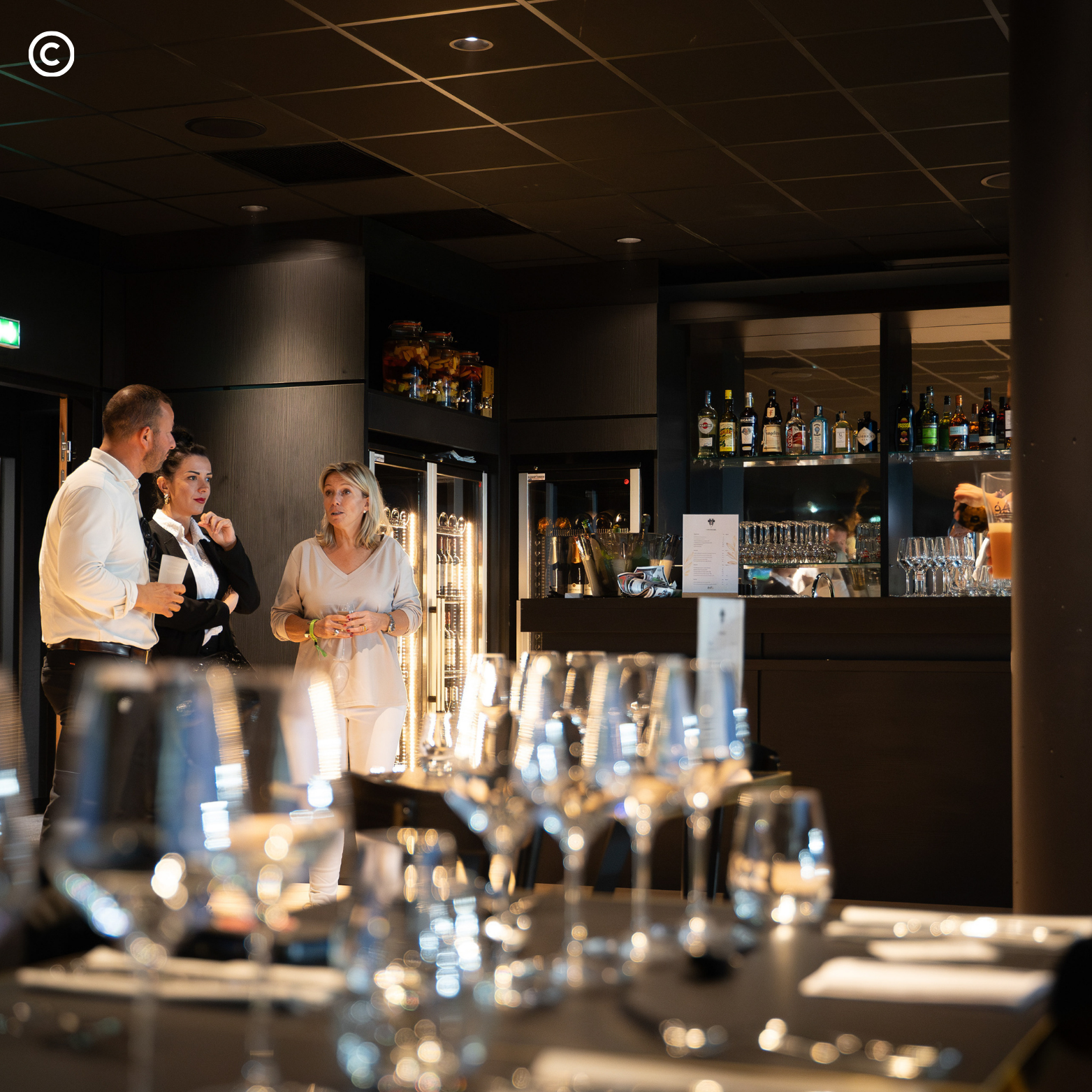 Trois personnes discutent au bar d'un restaurant ou d'un café élégant, avec des bouteilles d'alcool et des verres derrière elles.