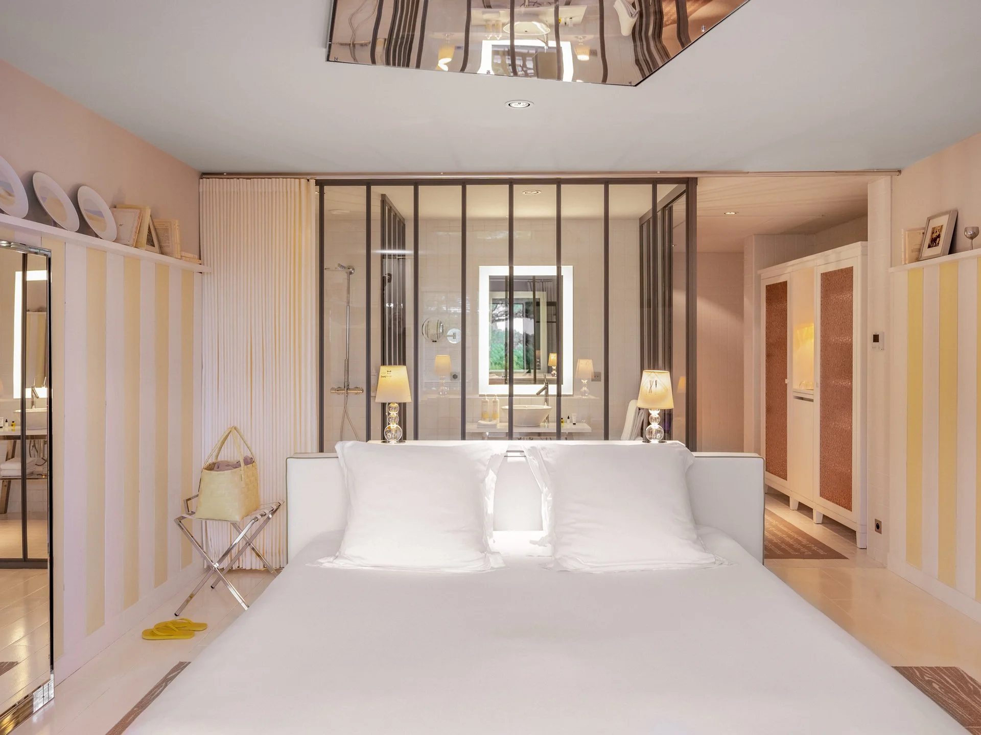Chambre avec lit blanc, ambiance lumineuse, décoration moderne avec lampes, miroirs, et salle de bain visible à travers une cloison en verre.