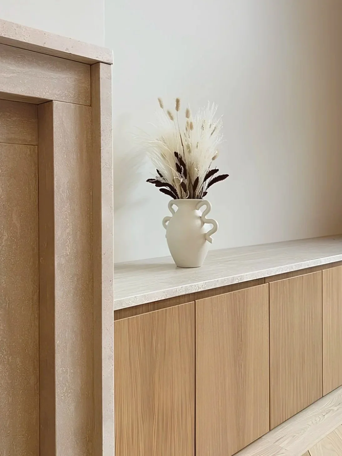 Vase blanc avec décoration de fleurs séchées blanches et brunes sur un meuble en bois clair.