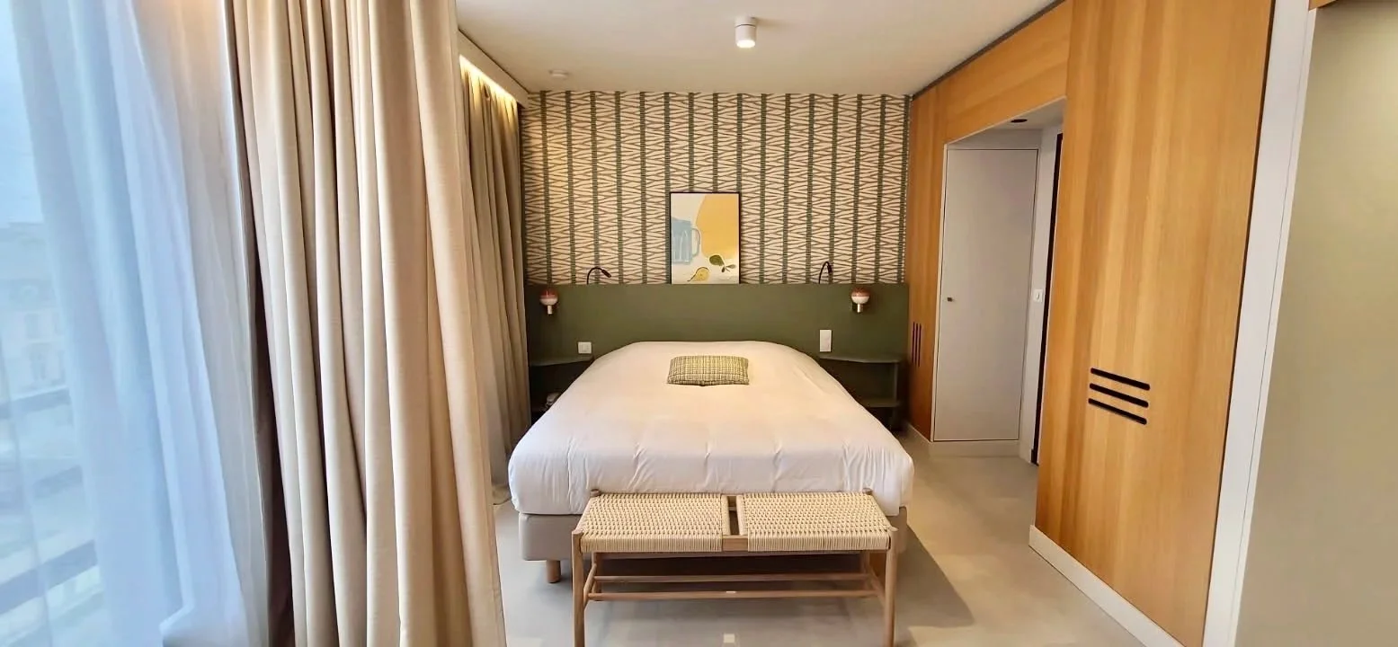 Chambre d'hôtel avec lit double, tête de lit verte, murs décorés avec un motif géométrique, une peinture abstraite, un banc en bois à la tête du lit, rideaux clairs et murs en bois.