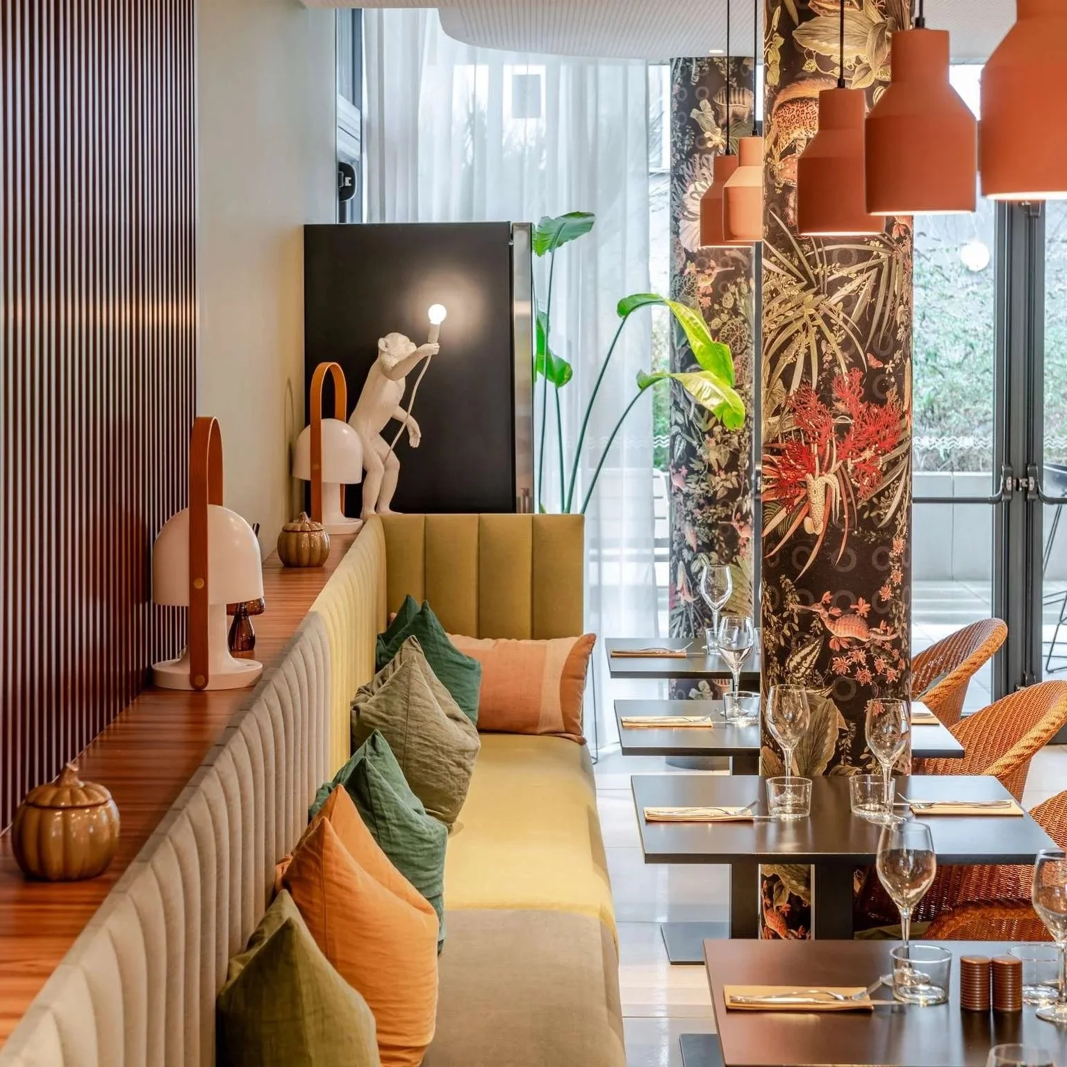 Intérieur d'un restaurant avec une banquette rembourrée, des coussins verts et orange, des tablettes avec lampes design, des plantes vertes, et des tables dressées avec des verres à vin et assiettes, avec une décoration florale sur un pilier.