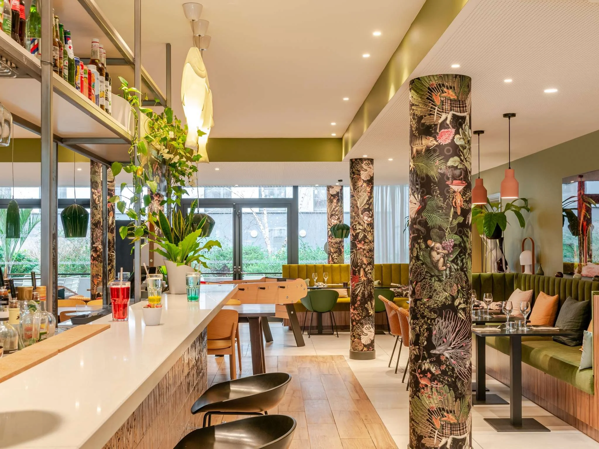 Intérieur d'un restaurant avec des colonnes recouvertes de tissu à motifs floraux et de jungle, un comptoir blanc avec des verres colorés, des plantes vertes et un éclairage moderne.