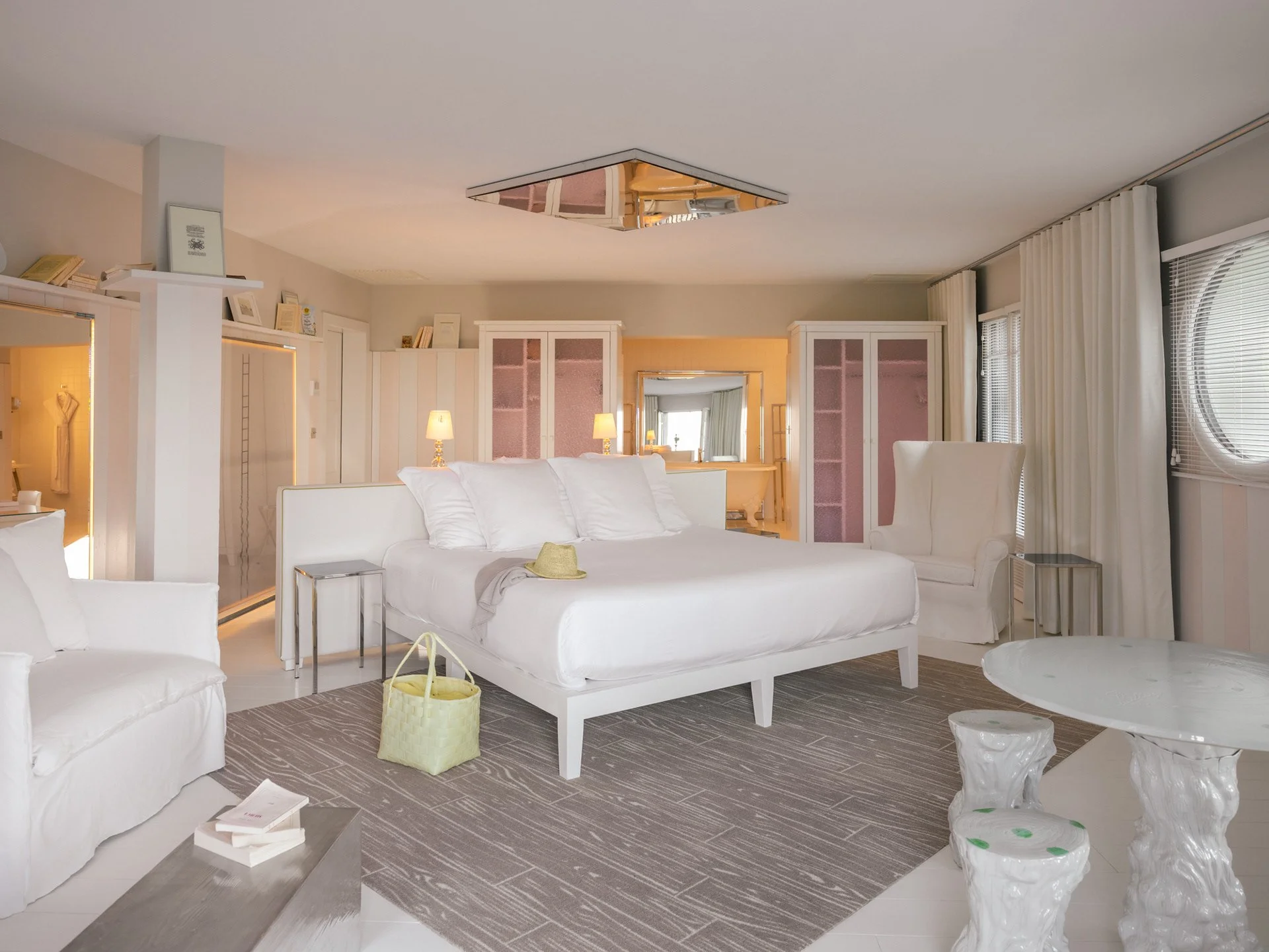 Salon blanc avec canapé, fauteuil, table et lampes, vue vers un miroir au plafond, fenêtre avec volets et rideaux.