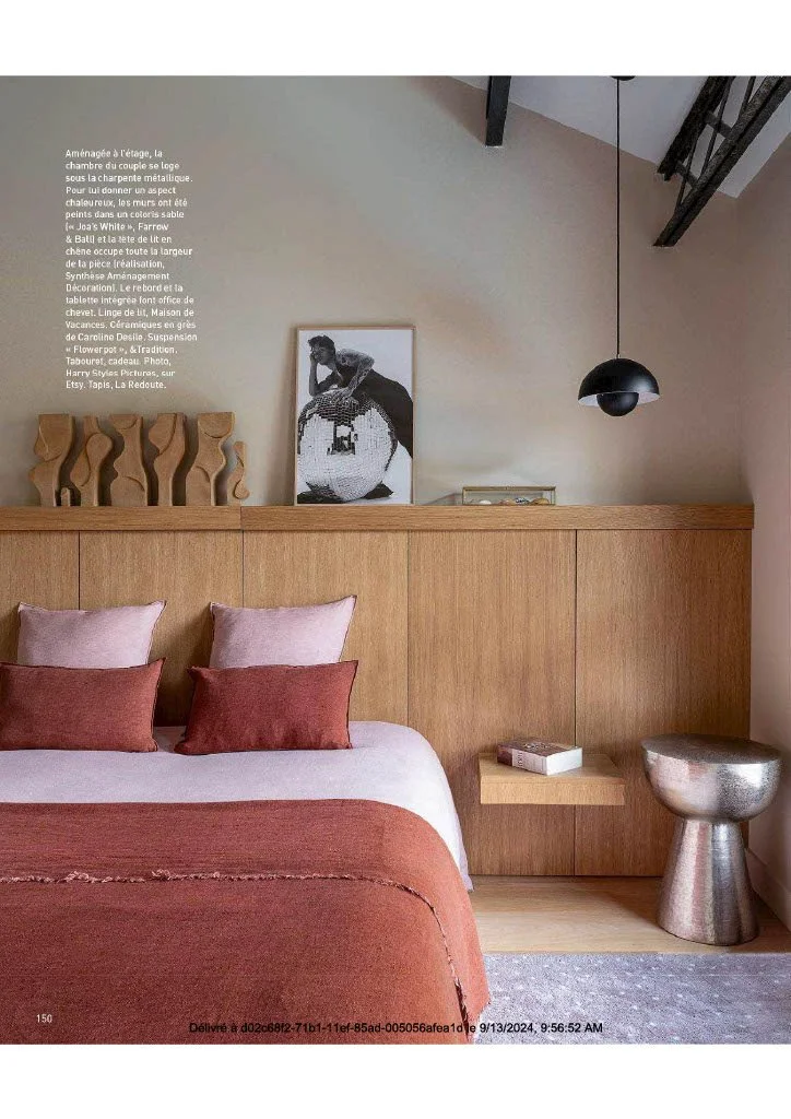 Chambre décorée avec lit avec oreillers roses et rouges, tête de lit en bois, table d'appoint en bois avec livre, lampe noire suspendue, cadre avec photo noire et blanche, décoration en bois sculpté, repose-pieds en métal, mur beige avec texte en fra