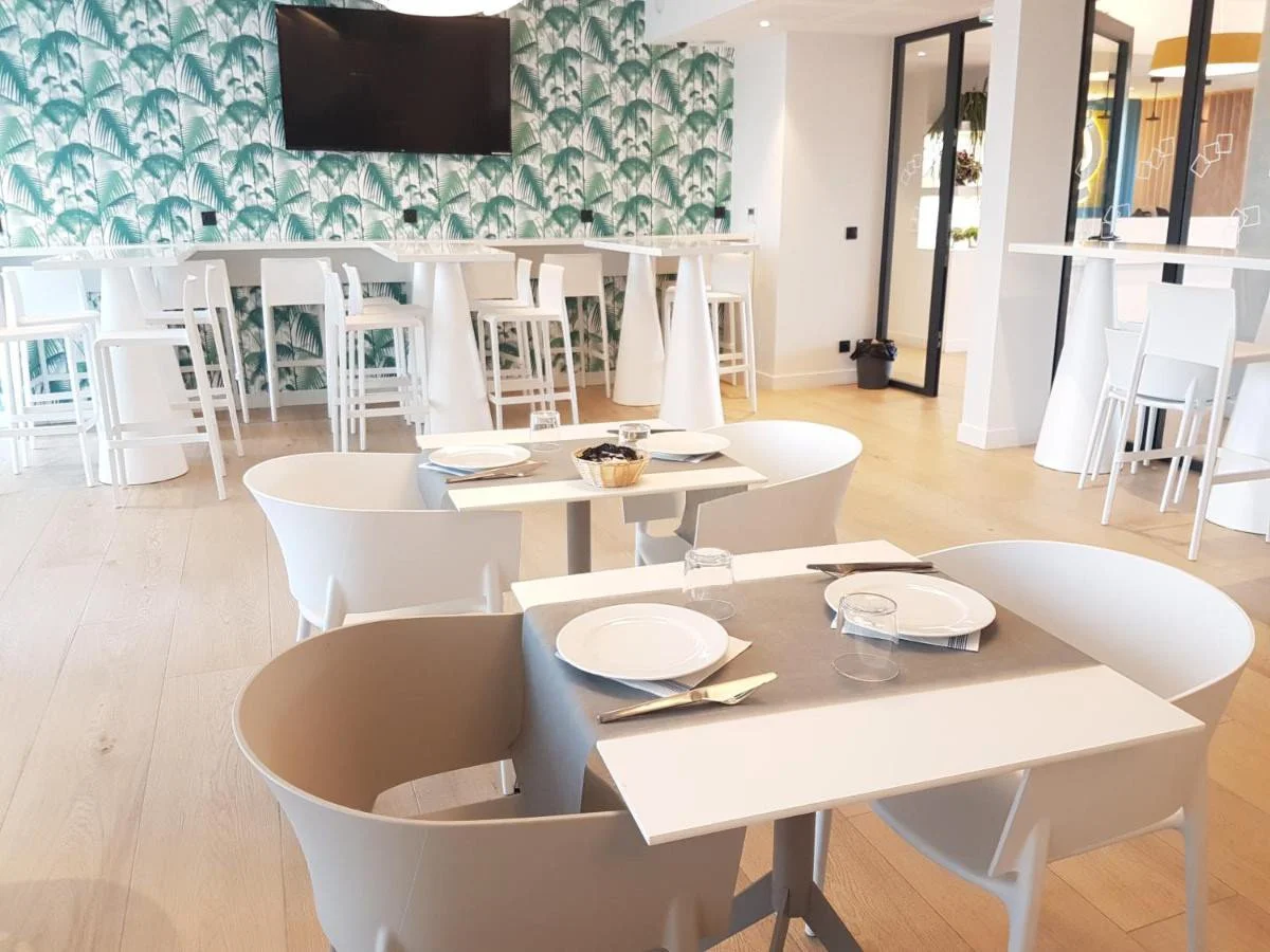 Salle à manger de restaurant avec tables et chaises blanches, décor vert tropical sur le mur, télévision, porte-fenêtre vitrée, et tables dressées avec assiettes, verres et couverts.